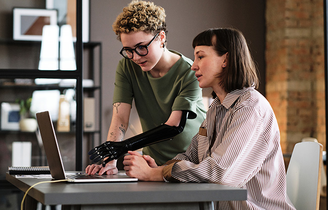 Young woman with prosthetic arm pointing at laptop and talking to her colleague during work at office